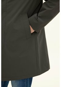 Mörkgrön regnrock med rak skärning, med sidofickor och en slät, glänsande yta. Ärmarna har något uppvikta manschetter.