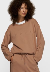 Sweatshirt beige à coupe décontractée, avec "10 DAYS" imprimé dans une teinte plus foncée. Texture douce et poignets côtelés, accompagné d'un pantalon assorti.