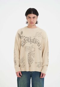 Beige Rippenpullover mit grafischen Drucken in Dunkelgrau, mit Rundhalsausschnitt und überschnittenen Schultern, kombiniert mit einer lässigen blauen Jeans.