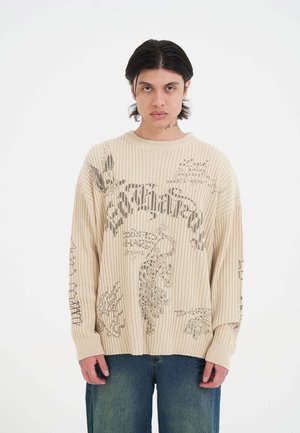 Beige Rippenpullover mit grafischen Drucken in Dunkelgrau, mit Rundhalsausschnitt und überschnittenen Schultern, kombiniert mit einer lässigen blauen Jeans.
