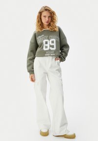 Overmaat groene sweatshirt met witte graphics, gecombineerd met losse witte cargo-broeken en chunky witte schoenen. Eenvoudige, casual streetwear-look.