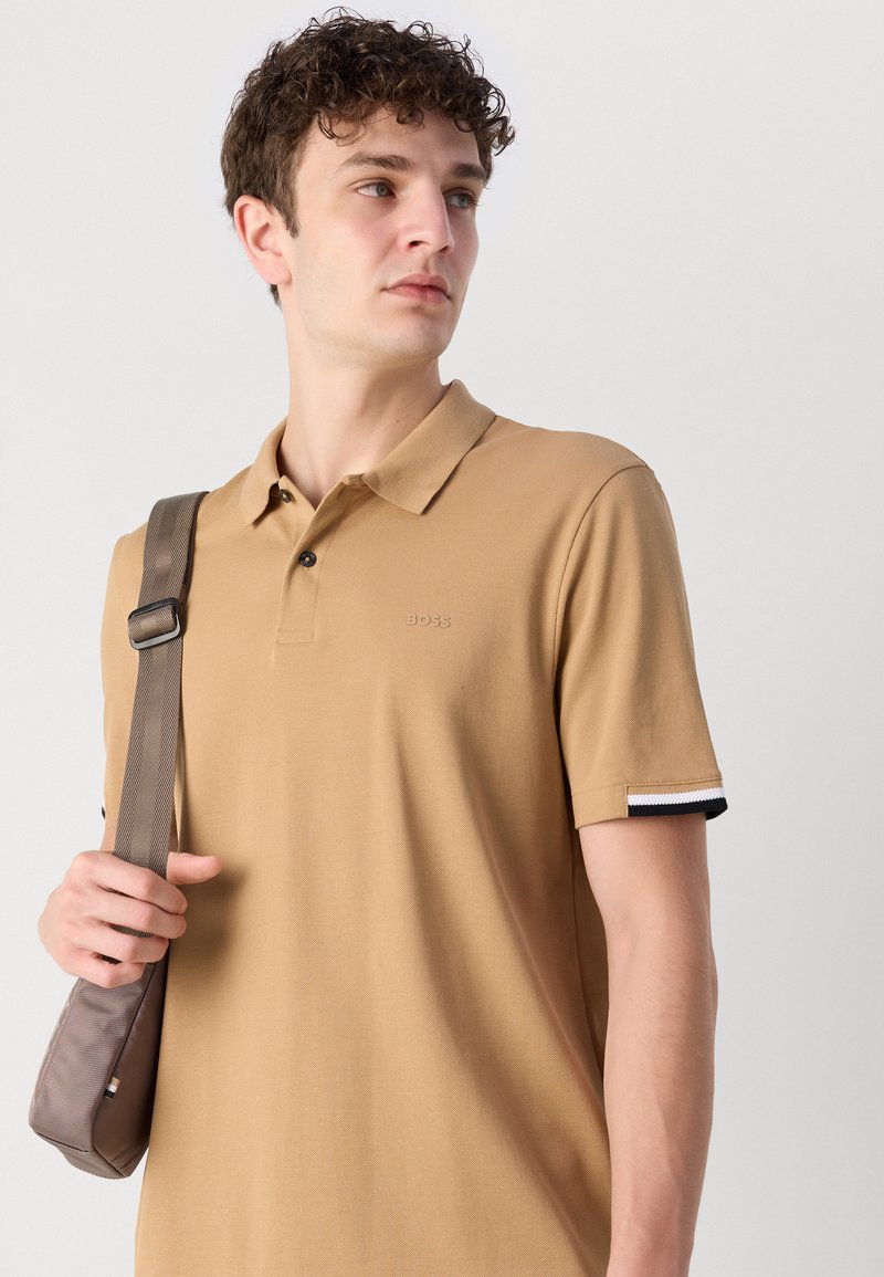 Polo shirt beige en mélange de coton avec un col classique, deux boutons noirs et des accents rayés noir et blanc sur les poignets.