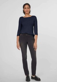 Marineblauer Strickpullover mit Bootsausschnitt und 3/4-Ärmeln. Dunkelgraue Slim-Fit-Jeans und schwarze Loafers mit Fellakzenten. Neutraler Hintergrund.