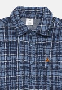 Blå rutete skjorte med knapper laget av mykt stoff, med krage, to blå knapper og en liten bjørne-logo brodert på brystlommen.