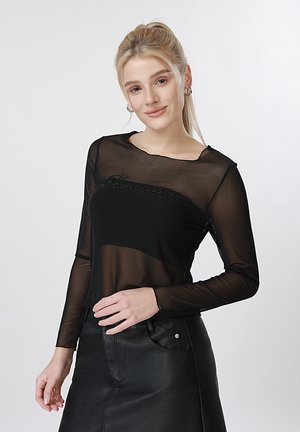 Jeune femme aux cheveux blonds portant un haut noir transparent à manches longues et une jupe en cuir noire, souriante avec la main sur la taille.