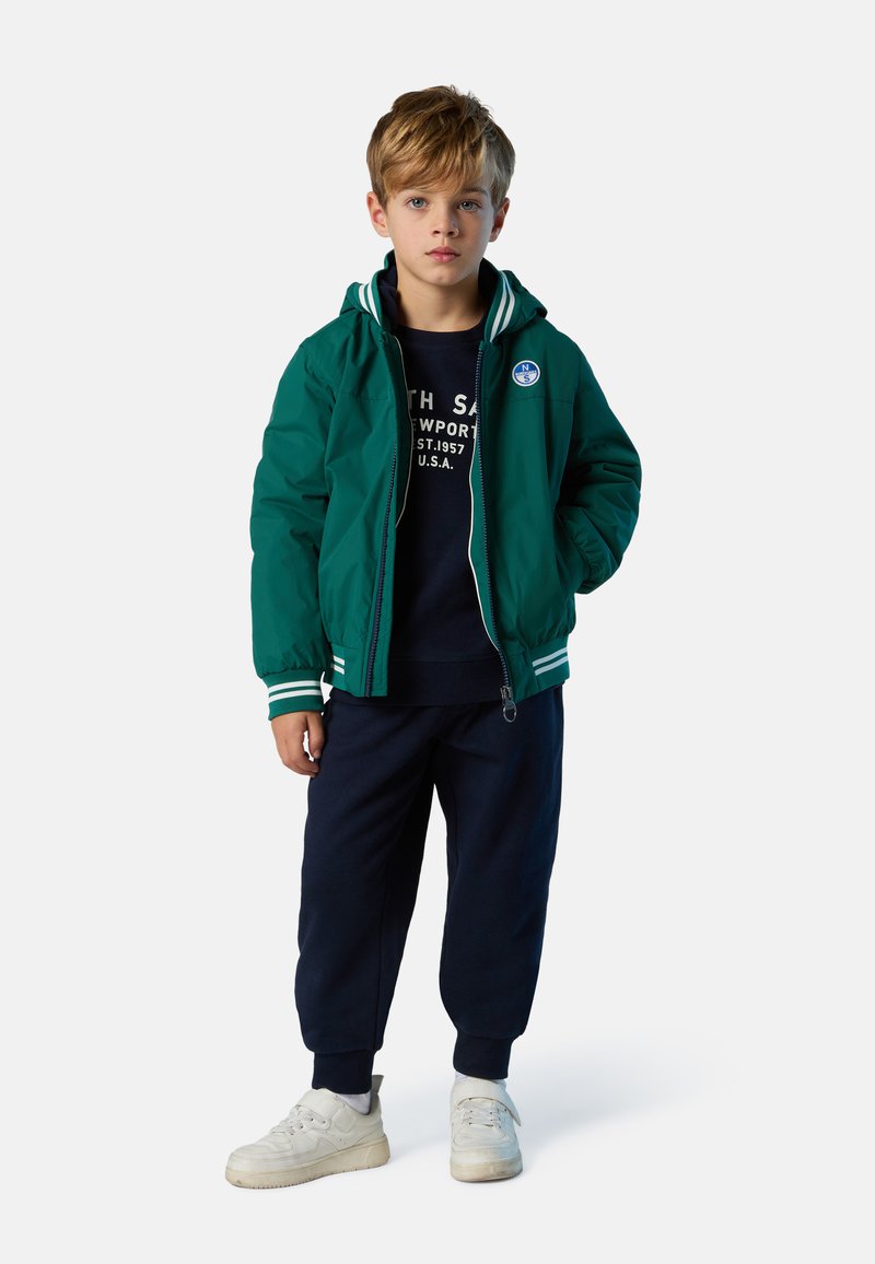 Giacca bomber verde con strisce bianche, zip frontale e cappuccio, indossata sopra una felpa blu scuro. Abbinata a pantaloni sportivi blu scuro e scarpe da ginnastica bianche.
