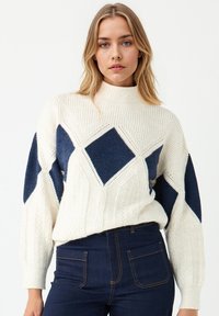 Kobieta z blond włosami, ubrana w biały sweter z dzianiny z dużymi granatowymi wzorami w romby oraz ciemnoniebieskie dżinsy o wysokim stanie na białym tle.