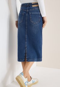 Femme portant une jupe midi en denim bleu avec des poches arrière et une fente au centre du dos, associée à des baskets blanches avec des accents bleus.