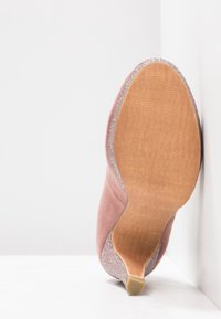 Chaussure à talon en daim rose unique avec talon pailleté et bordure, semelle tournée vers l'avant sur fond d'angle blanc.