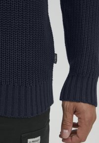 Maglione lavorato a maglia blu scuro con polsini e orlo a coste, caratterizzato da un motivo testurizzato e una piccola etichetta del marchio sul lato.