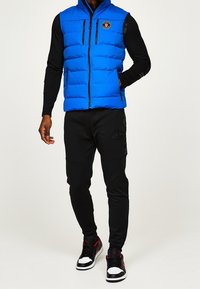 Blauer Puffer-Weste mit schwarzen Reißverschlussakzenten und Logo, kombiniert mit schwarzen Jogginghosen und schwarz-roten Sneakers. Glatte Textur, sportliche Passform.