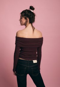 Bordeauxrotes Rippenoberteil mit Carmenausschnitt, kombiniert mit dunklen Jeans, zeigt lange Ärmel und eine figurbetonte Silhouette. Glatte Stofftextur.