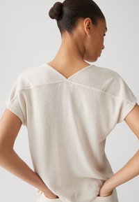 Top beige chiaro a maniche corte con un tessuto a maglia testurizzato e uno scollo a V sul retro. Presenta orli arrotolati e una vestibilità rilassata.
