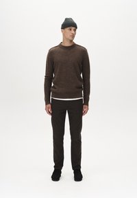 Bruine wollen trui met ronde hals, gedragen over een witte shirt. Donkerbruine op maat gemaakte broek en zwarte schoenen. Model staat tegen een witte achtergrond.