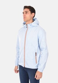 Giacca con cappuccio azzurro chiaro, con polsini elasticizzati, dotata di una cerniera frontale, dettagli arancioni e una tasca laterale con cerniera. Materiale liscio e leggero.
