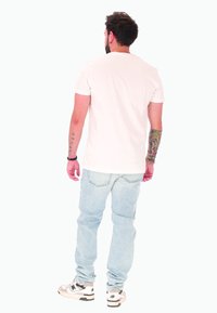 Uomo con tatuaggi che indossa una maglietta bianca, jeans azzurri chiari e sneaker bianche, in piedi di spalle su uno sfondo bianco uniforme.