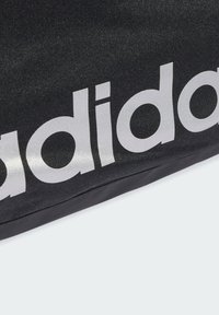 Musta kangaskassi, jossa on suuri, kiiltävä hopeinen "adidas"-logo. Pinnassa on kevyt kiilto, ja muoto on suorakulmainen, jossa on sileät reunan.
