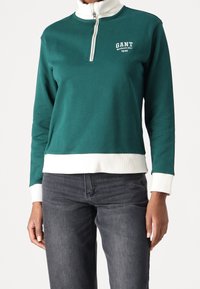 Sudadera verde con cremallera fabricada en algodón, con cuello y puños blancos. Presenta un logotipo impreso y un dobladillo acanalado. Diseño ajustado.