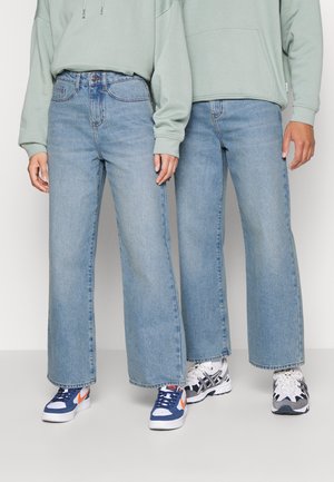 Deux personnes se tenant côte à côte, portant des jeans larges bleu clair, des hoodies vert clair et des baskets de sport avec des détails blancs, navy et orange.