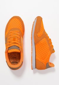 Klart orange sneakers med beige detaljer och grå sulor, med ett mesh-övre och snören, visade på en vit bakgrund.