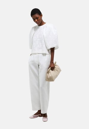 Femme portant un haut blanc texturé à manches bouffantes, un pantalon large blanc, tenant une pochette beige froncée, et portant des ballerines rose clair.