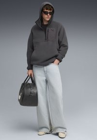 Grau gefütterter Pullover mit einem halben Reißverschluss, Fronttasche und Kapuze; kombiniert mit hellblauen, weit geschnittenen Jeans. Schwarze Lederhandtasche mit Griff.