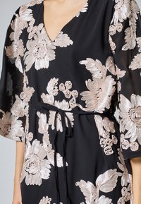Klänning med svart tyg och stora blommönster i ljusrosa, V-neckline och ett självknutet bälte i midjan. Fladdrande ärmar.