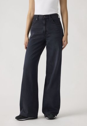 Oseba, oblečena v temne kavbojke z visokim pasom in širokimi hlačnicami, belo brezrokavnik in črne superge na enobarvnem svetlo sivem ozadju.