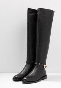 Botas de cuero negro hasta la rodilla con un acabado texturizado, que presentan una correa con tachuelas y una hebilla en tono dorado. Diseño de punta redonda y tacón bajo.