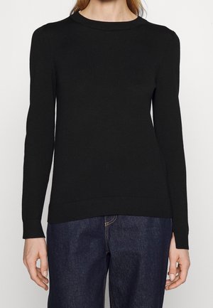 Femme portant un pull noir uni à manches longues et un jean bleu foncé, debout devant un fond neutre.
