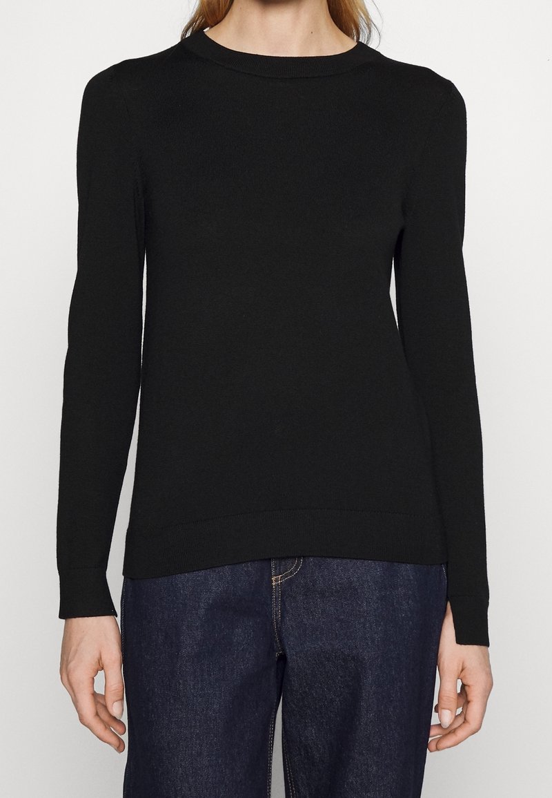 Femme portant un pull noir uni à manches longues et un jean bleu foncé, debout devant un fond neutre.
