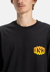 Jongere in een zwart T-shirt met feloranje "DGSD" logo op de linkerborst, tegen een effen lichte achtergrond.