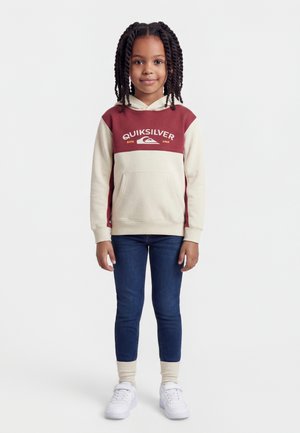 Nuori tyttö seisoo, pukeutuneena kaksiväriseen punaisen ja kermanväriseen Quiksilver-huppariin, tummansinisiin farkkuihin, kermanvärisiin sukkiin ja valkoisiin tennareihin.