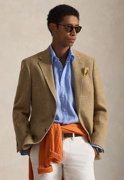 Μπεζ σακάκι με σχέδιο houndstooth, ανοιχτό μπλε πουκάμισο με κουμπιά, πορτοκαλί πουλόβερ δεμένο στη μέση, λευκό παντελόνι και σκούρα γυαλιά ηλίου. Υφασμάτινη υφή.