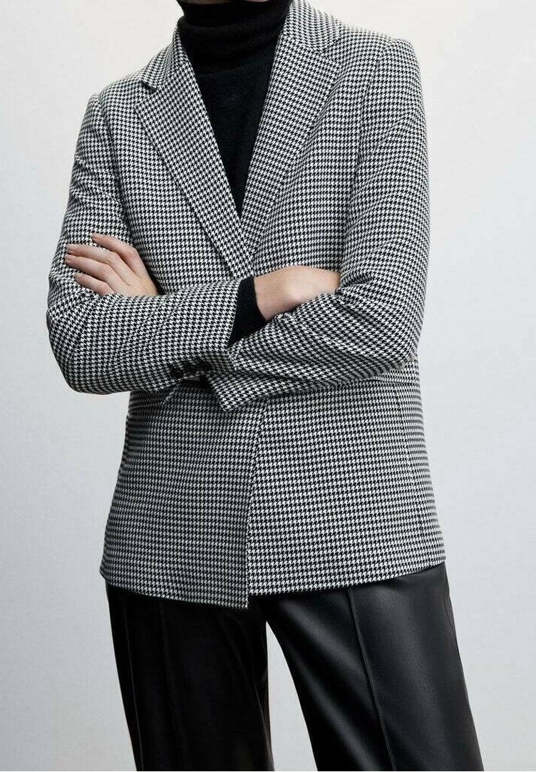 Personne portant un blazer à motif pied-de-poule noir et blanc, un col roulé noir et un pantalon en cuir noir, les bras croisés devant un fond uni.