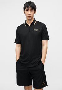 Μαύρο πόλο πουκάμισο με φερμουάρ μέχρι τη μέση, χρυσές λεπτομέρειες και λογότυπο. Συνδυάζεται με μαύρο σορτς με υφή. Απλός σχεδιασμός, casual εφαρμογή.