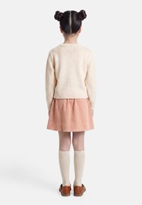 Enfant aux cheveux bruns coiffés en deux chignons, portant un pull crème, une jupe rose, des chaussettes crème montant jusqu'aux genoux et des chaussures marron, debout de dos.