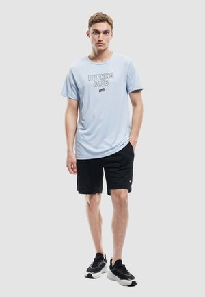 T-shirt de sport bleu clair avec un graphisme "RUNNING CLUB", associé à un short noir. Le modèle porte des chaussures de course noires et blanches.