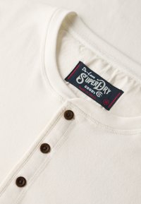 Helle Baumwollbluse in Cremefarbe mit Rundhals, Knopfleiste mit braunen Knöpfen und einem Etikett mit "Superdry"-Markendetails.