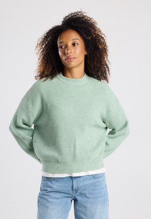 Mintgroene gebreide trui met ribbelstructuur, ronde hals en verlaagde schouders, gecombineerd met lichtblauwe spijkerbroek.