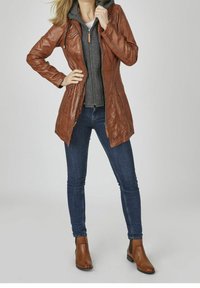 Giacca di pelle marrone con zip e cappuccio, sovrapposta a un felpa grigia. Abbinata a jeans blu e stivaletti marroni.