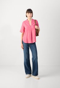 Camicia di lino rosa con scollo a V, maniche corte e vestibilità rilassata, abbinata a jeans in denim blu a zampa d'elefante e sneakers bianche. Inclusa borsa a spalla marrone.