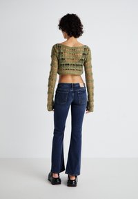 Grønn åpenstrikket cropped genser med lange ermer, kombinert med mørkeblå flared jeans, satt mot en nøytral bakgrunn.