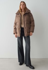 Giubbotto puffer marrone con un collare in finto pelliccia strutturato. Presenta una chiusura lampo frontale e tasche laterali, abbinato a jeans larghi scuri.
