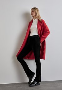 Cappotto rosso di lana sopra un dolcevita bianco, abbinato a pantaloni a zampa d'elefante in velluto a coste neri e scarpe stringate nere, su uno sfondo chiaro.