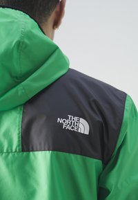 Osoba nosi zelenu i crnu jaknu s kapuljačom na kojoj je vidljiv logo The North Face na gornjem dijelu leđa.