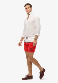 Chemise en lin blanche à manches longues, short de bain rouge avec l’imprimé 'SUPER DRY CO.', et chaussures de bateau marron. Tenue d'été décontractée.