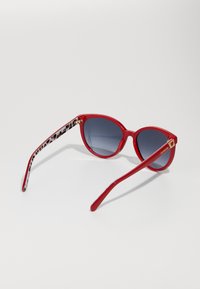 Κόκκινα γυαλιά ηλίου σε σχήμα γάτας, με σκουρόχρωμα φακούς σε βαθμίδες και βραχίονες με ασπρόμαυρο σχέδιο που διαθέτουν χρυσή λεπτομέρεια στη στήριξη.