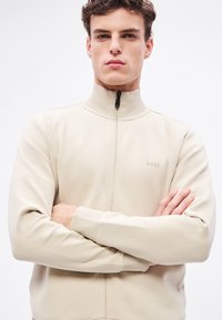 Bézs zipzáras dzseki magas gallérral, puha anyagból készült. Bordázott mandzsettákkal és a mellkason lágyan hímzett márkajelzéssel rendelkezik.