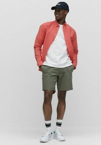 Rode button-up shirt, witte t-shirt, olijfgroene shorts en witte sneakers met groene accenten. Zwarte pet met tekst. Effen achtergrond.